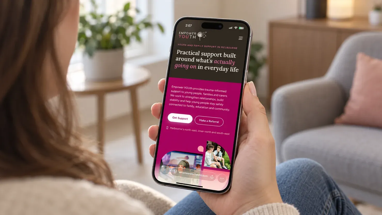 The Empower YOUth home page on a phone, held in one hand in a living room: the headline, the Get Support and Make a Referral buttons, and the start of the photographs below them.